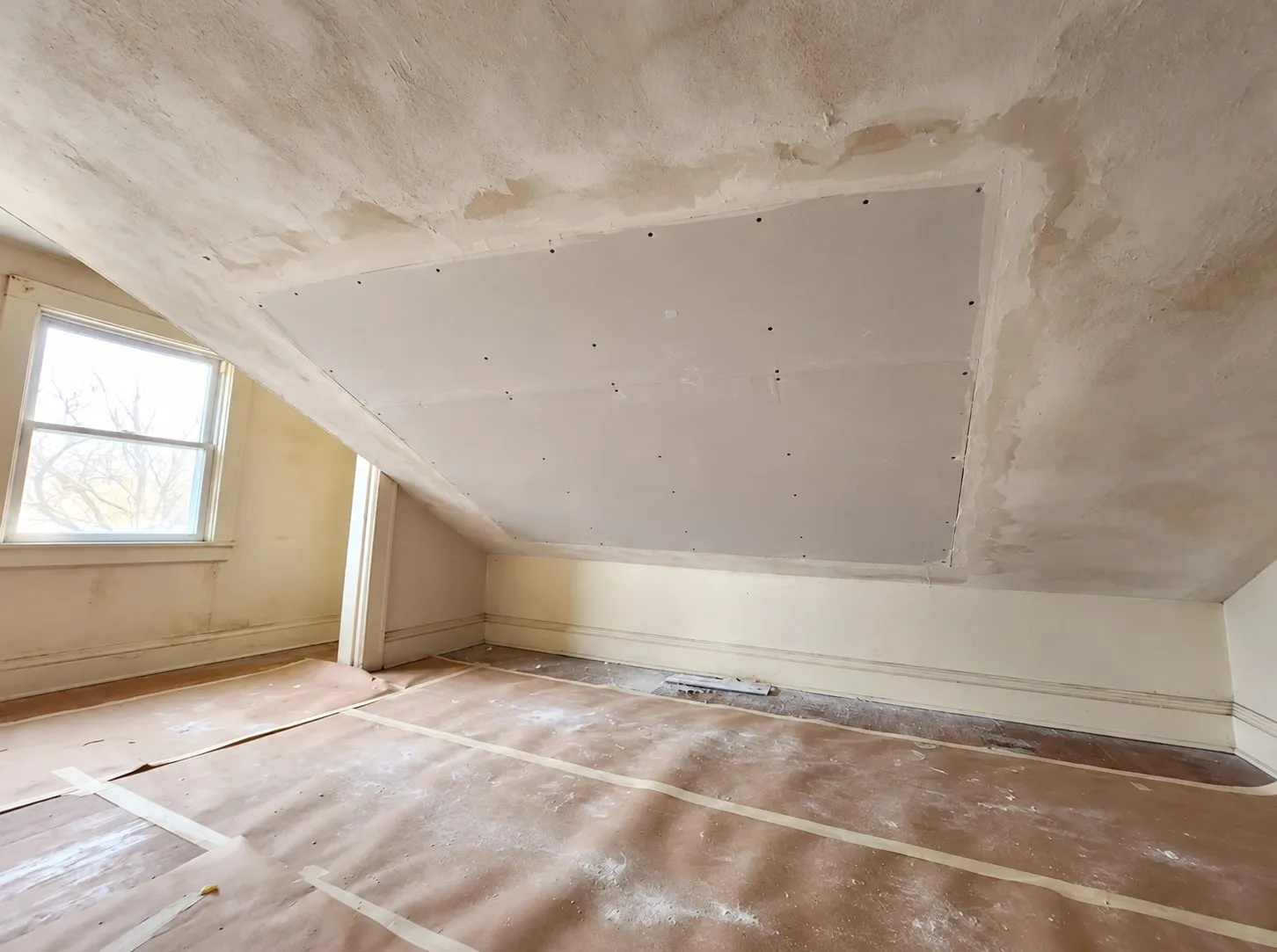 Water-stained sloped ceiling with the damaged section replaced by new drywall, screwed in and ready for taping, floor covered in paper