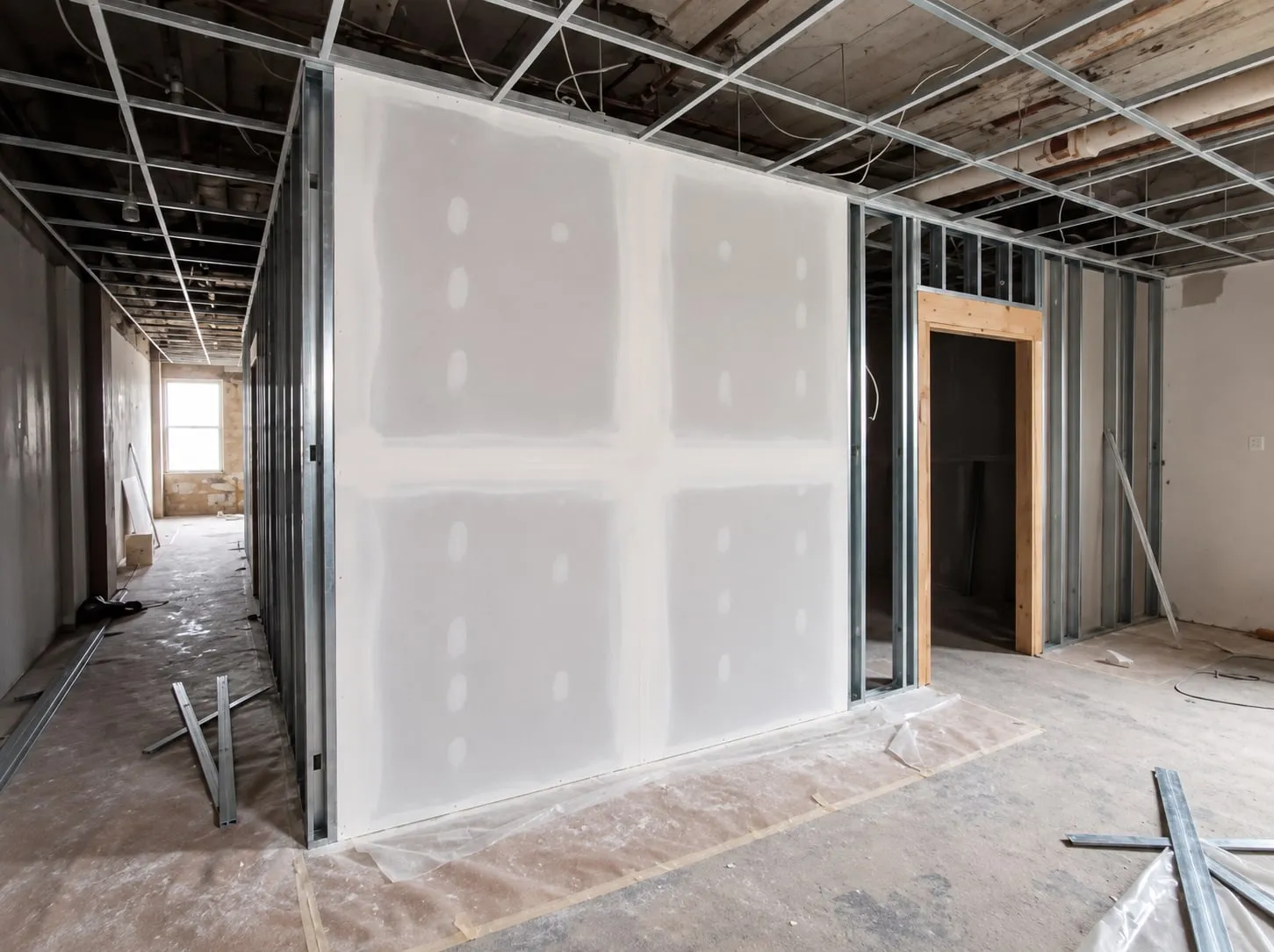Drywall partition on metal studs in a gutted commercial floor, seams taped and first-coated, with open stud framing and a door opening beside it