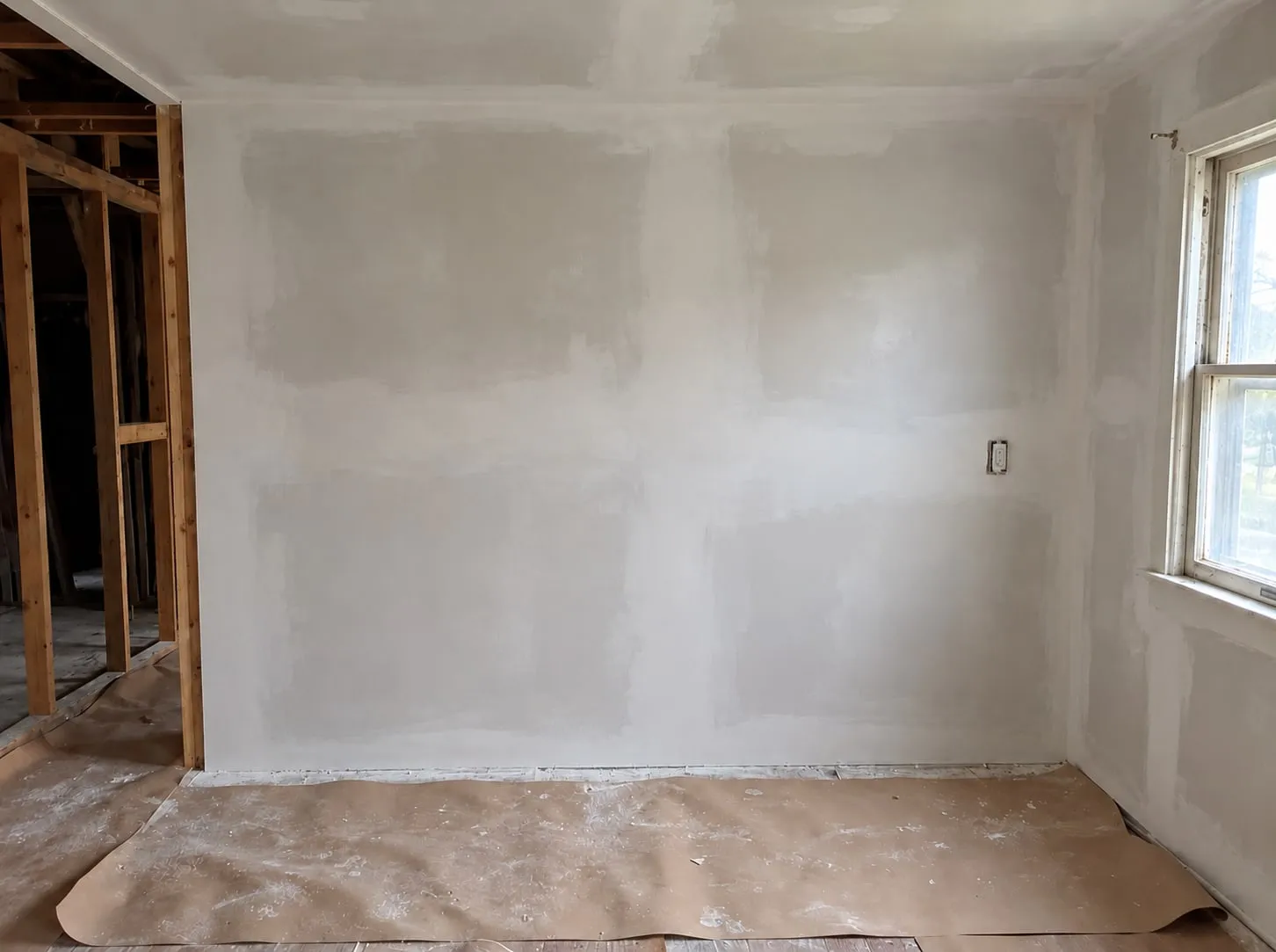 Room remodel in progress: a wall and ceiling skim-coated in patches beside open stud framing, with the floor covered in paper