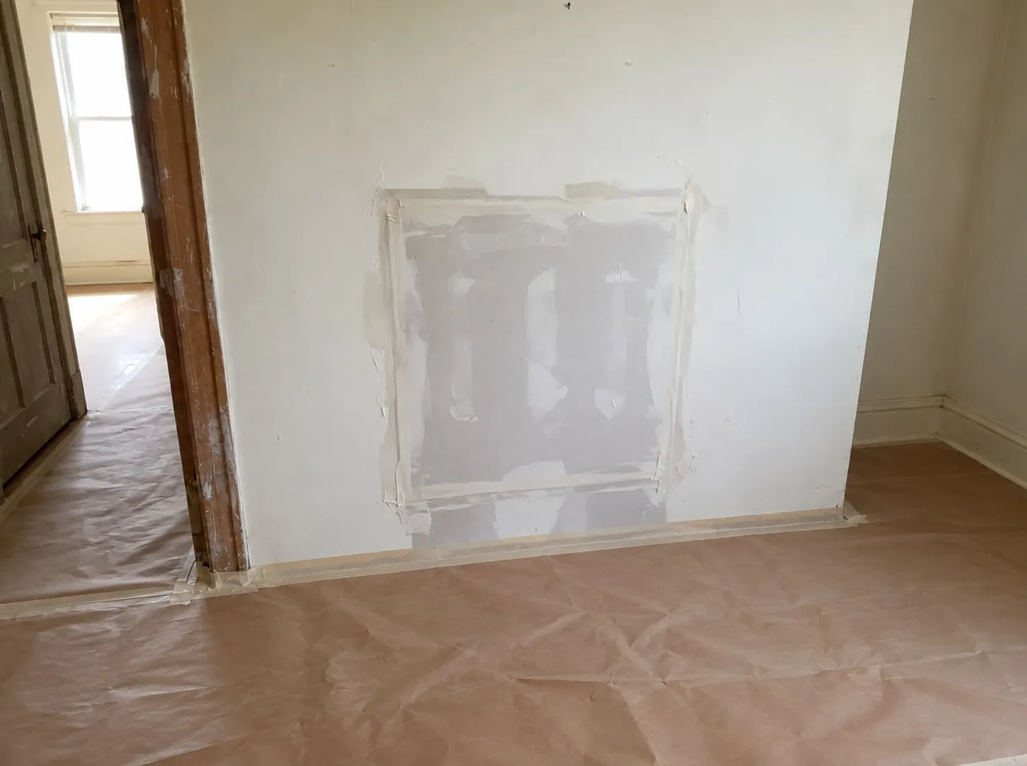 Square drywall patch in a wall beside a doorway, seams taped and first-coated, with paper protecting the wood floor